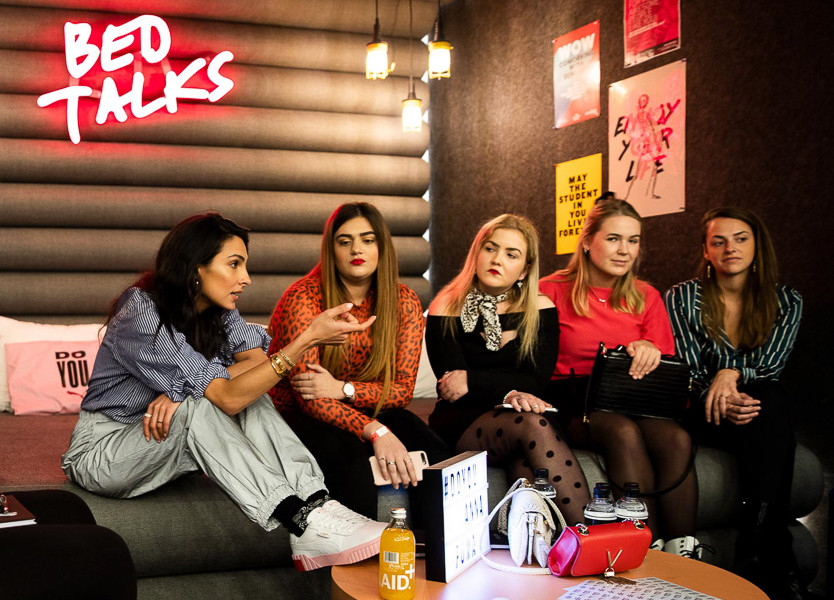 Five girls talk on a bed beneath a 'BedTalks' neon sign on The Student Hotel Maastricht